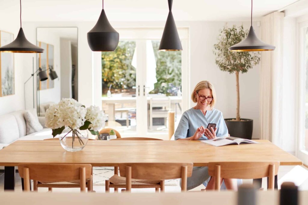 Senior Woman Relaxing With Magazine At Home Looking At Mobile Phone Sitting At Dining Room Table