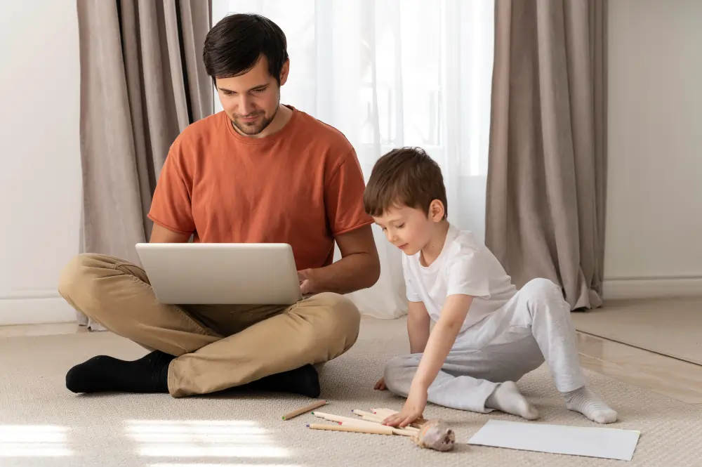 A father sits cross-legged on the floor working on a laptop while his young son draws beside him, depicting shared space during remote work.